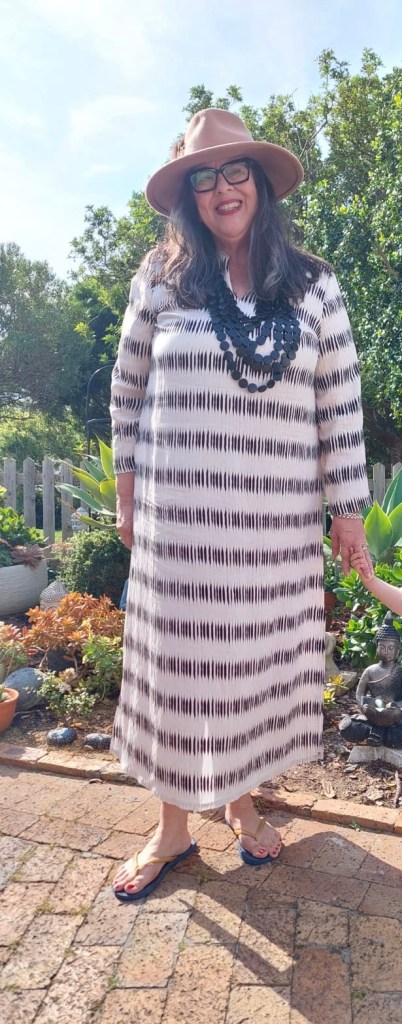 A woman wearing a patterned maxi dress and a wide-brimmed hat, smiling in a garden setting with greenery and potted plants in the background.