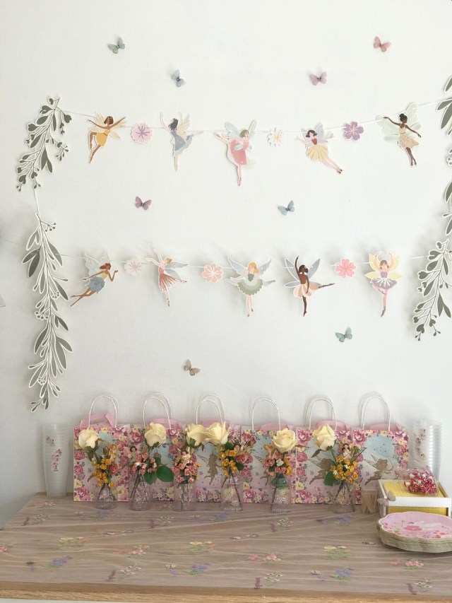 A decorated wall featuring paper ballerina cutouts and floral designs, with a wooden table adorned with small glass vases of flowers and decorative gift bags.