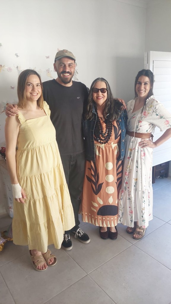 A group of four people posing together in a bright room. Two women on the left wear casual dresses, one in yellow and the other in a floral design. A man in the center is wearing a black t-shirt and cap, and a woman on the right is dressed in a patterned long dress. The room has light-colored tiles and a simple white decor.