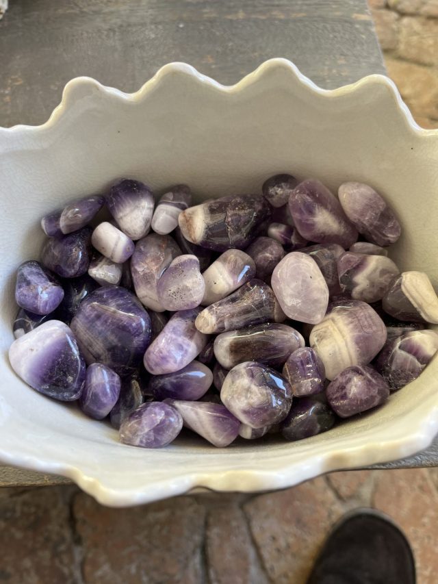 A white ceramic bowl filled with various polished purple stones, including amethyst, showing different shapes and shades of purple.