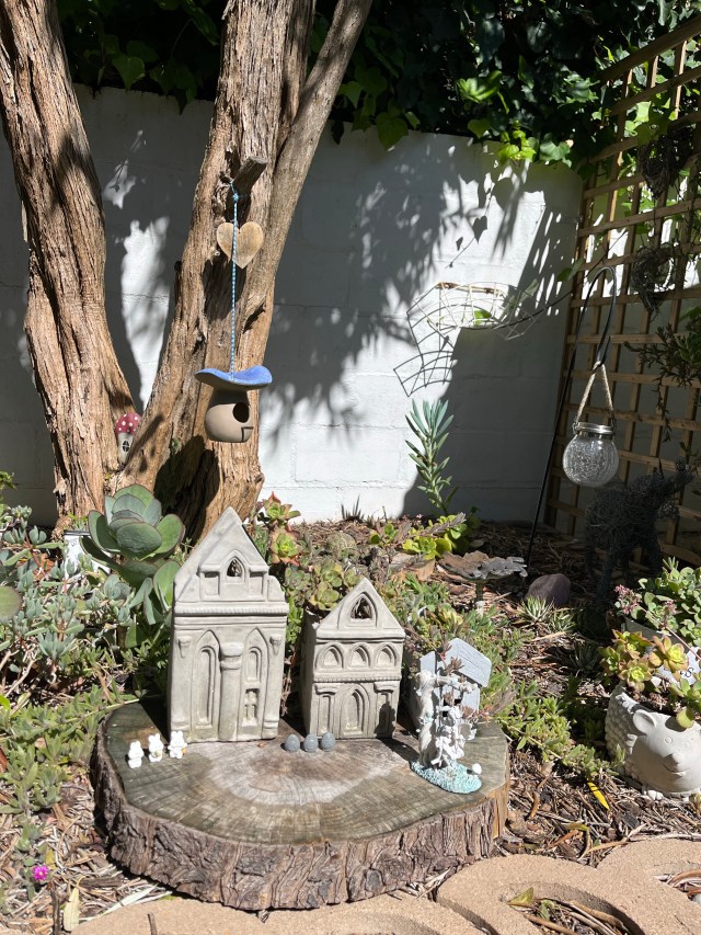 A small garden scene featuring two decorative concrete houses on a tree stump, surrounded by succulents. A birdhouse and heart-shaped decor hang from a tree, with a glass lantern and various garden decorations visible in the background.