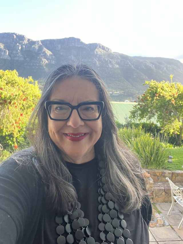 A woman with long dark hair and glasses smiles for a selfie, wearing a black top and a large beaded necklace. In the background, a scenic view of mountains and greenery is visible.