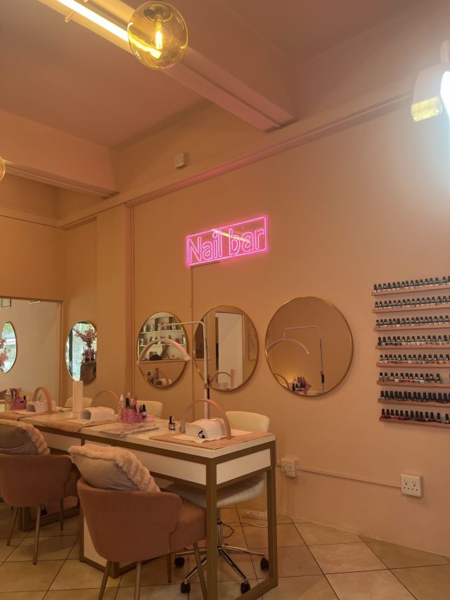 Interior of a nail bar featuring a pink neon sign, elegant seating, and a display of nail polish options on the wall.