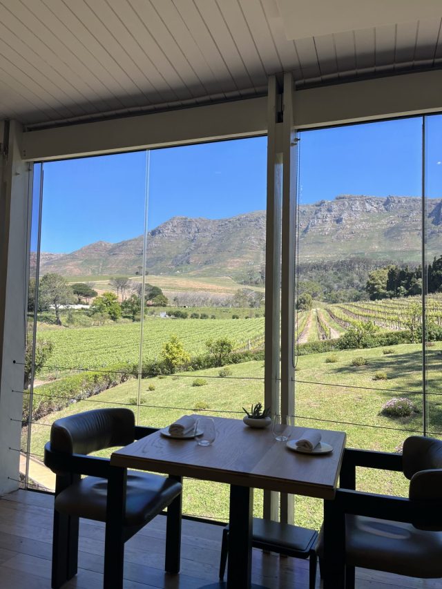 A view from inside a restaurant overlooking a vineyard and mountains under a clear blue sky, with a dining table set for two.