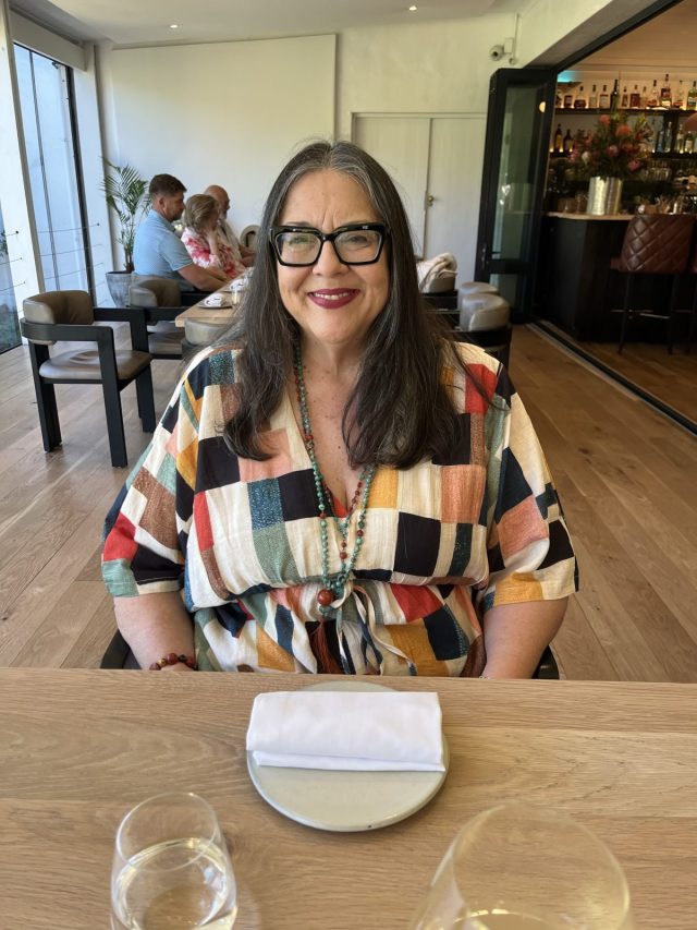 A woman with long hair and glasses sits at a dining table, wearing a colorful patterned shirt. In front of her is a table setting with a plate and napkin. The background shows a relaxed restaurant interior with people dining.