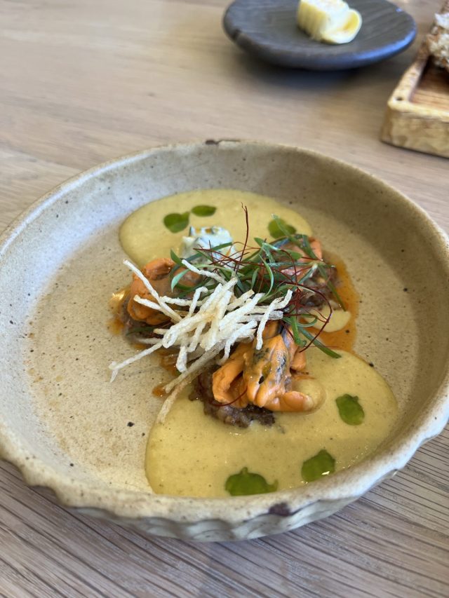 A gourmet dish featuring creamy yellow sauce, topped with sautéed shrimp, microgreens, and crispy fried noodles, served in a rustic bowl.