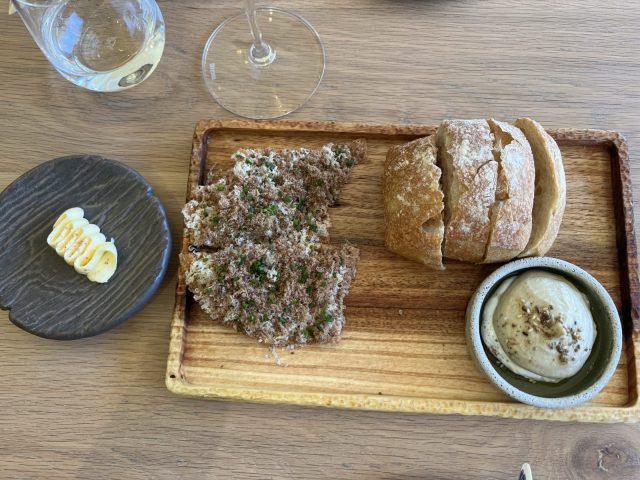 A wooden platter featuring artisan bread, a piece of crispy topping with herbs, and a small bowl of creamy dip, accompanied by a decorative black plate with a piece of corn on the side.