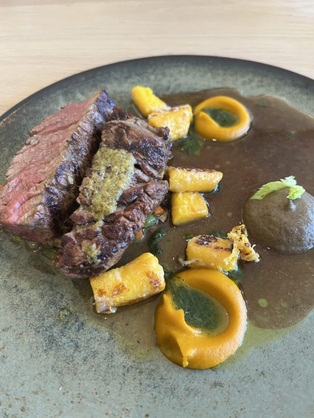 A plate featuring sliced beef with a herb sauce, accompanied by colorful root vegetable puree, sautéed pieces of yellow squash, and garnished with green sauce.