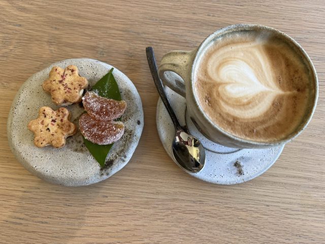 A cup of latte art coffee beside a plate of assorted cookies, including flower-shaped and fruit-shaped treats, on a wooden table.