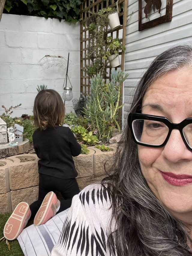A woman with long, gray hair and glasses smiles at the camera while a young child in black clothes tends to plants in a garden behind her.