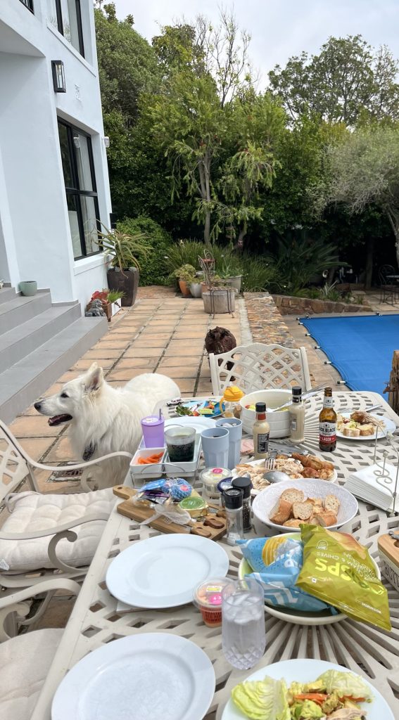 An outdoor dining scene featuring a white dog resting beside a table filled with various foods and drinks, including plates, snacks, and beverages. The setting has a backdrop of lush greenery and a blue pool.