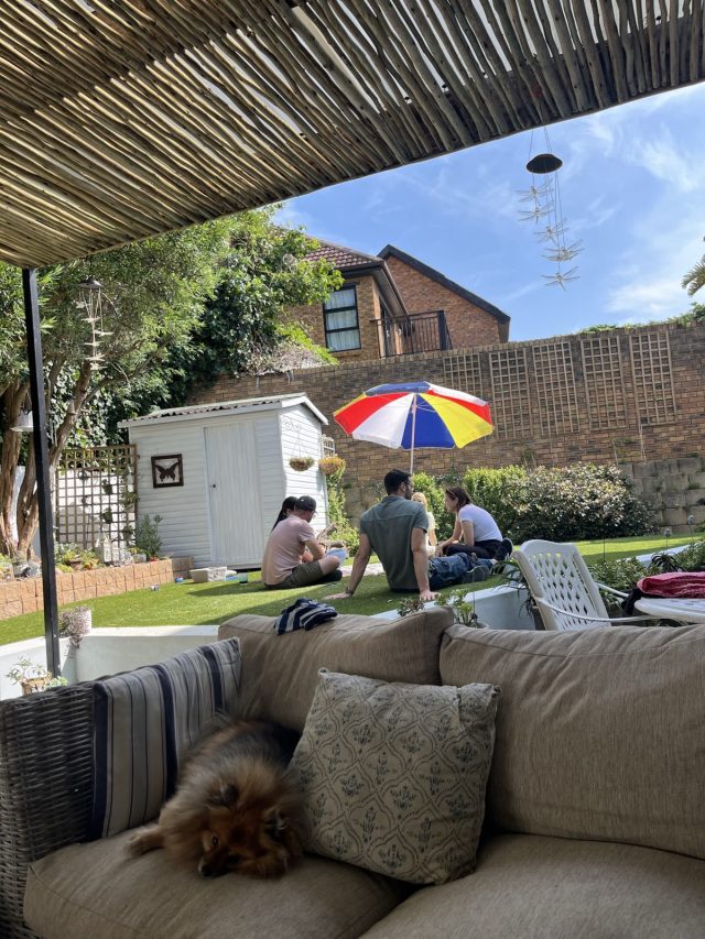 A view of a backyard with a sofa in the foreground, featuring a sleeping dog, and three people sitting on the grass under a colorful umbrella. A white shed and greenery are visible in the background.