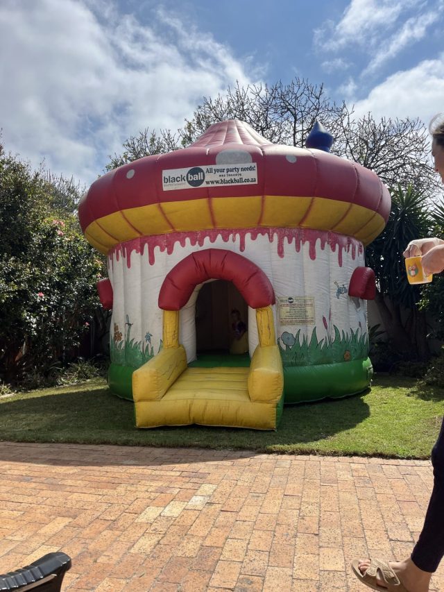 A colorful, inflatable bounce house designed like a mushroom, featuring a yellow and red roof, with a grassy area surrounding it and a person in the foreground holding a drink.
