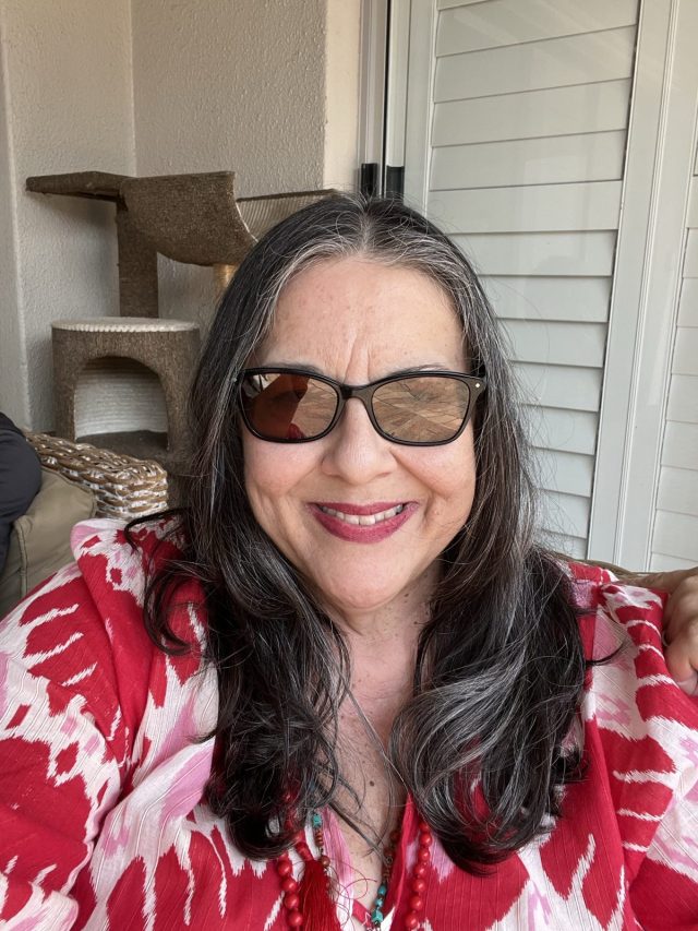 A woman with long, wavy hair and sunglasses smiles while sitting indoors. She is wearing a red and white patterned top and has a casual, happy demeanor.