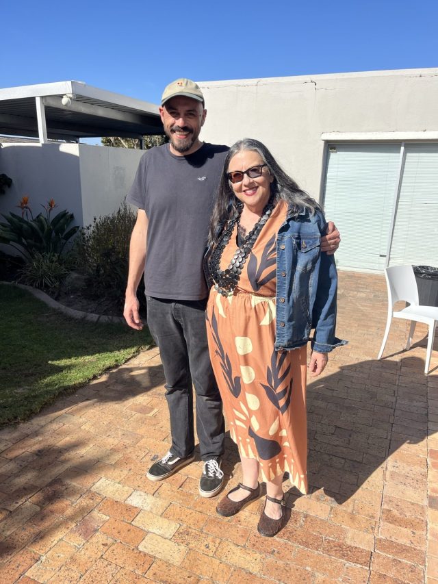 A man and a woman are standing together outdoors. The man is wearing a gray t-shirt and a cap, while the woman is dressed in a patterned orange dress with a denim jacket. They are smiling and posed on a brick pathway, with greenery and a building in the background under a clear blue sky.