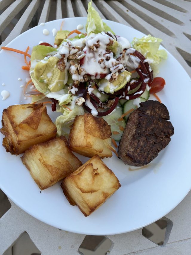 A plate of food featuring a portion of grilled meat, crispy potato cubes, and a fresh salad with lettuce, avocado, and drizzled dressing.