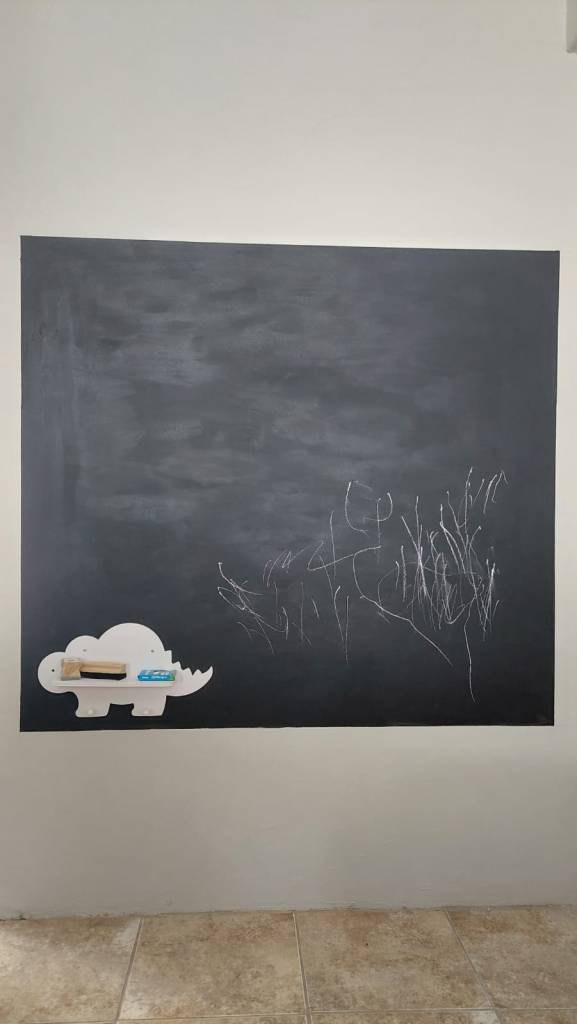 A large black chalkboard featuring white chalk markings and a wooden dinosaur-shaped chalk holder with chalk and an eraser.